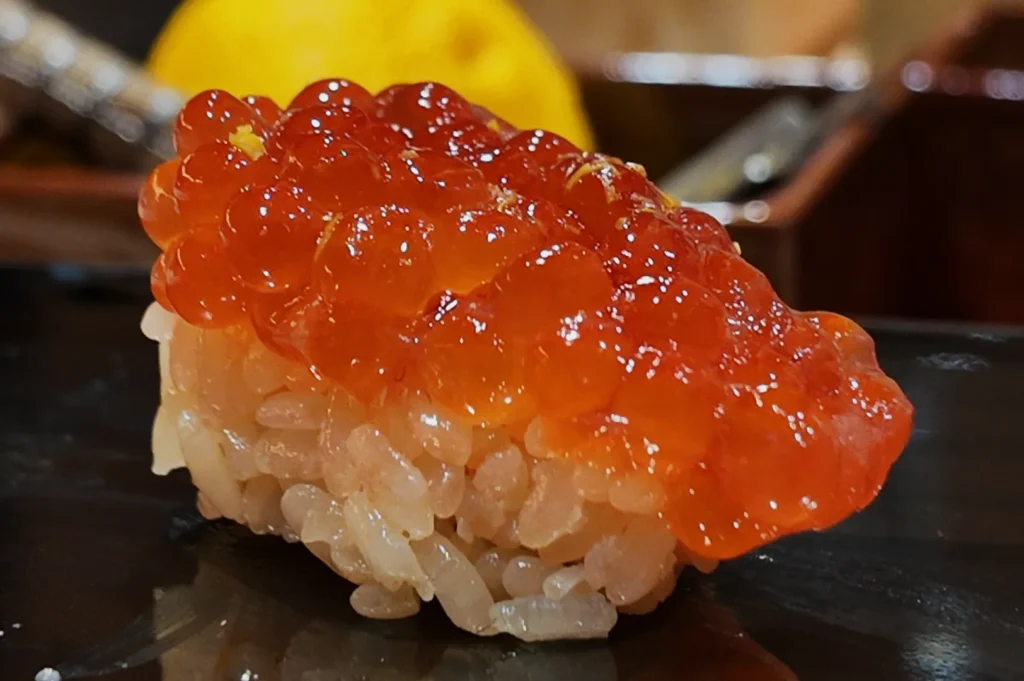 A piece of sushi topped with colorful fish roe placed on a black plate. A lemon placed in a wooden box can be seen in the background.