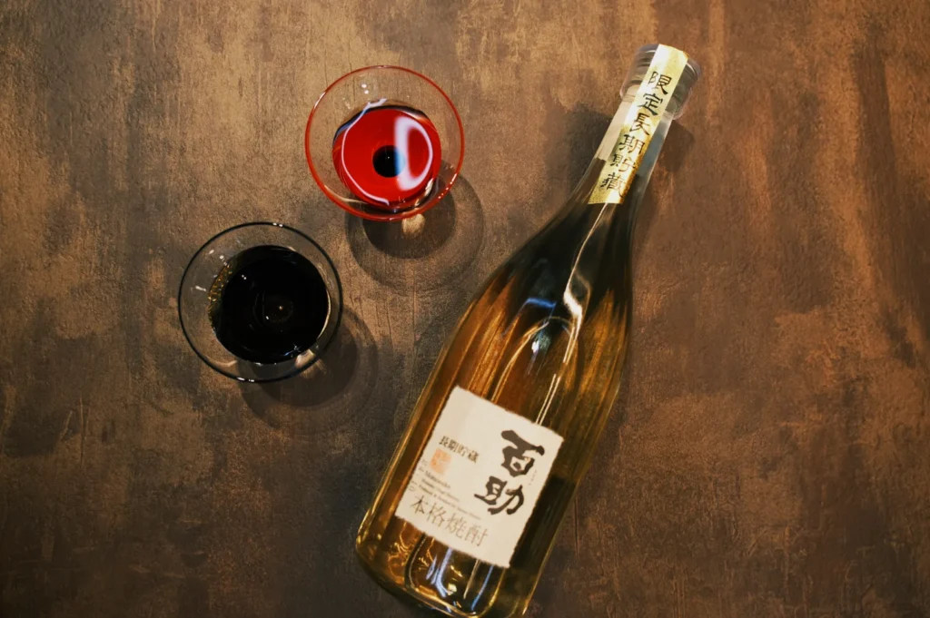 A bottle of sake with two glasses, one filled with a red liquid and the other with a dark liquid, arranged on a wooden surface.