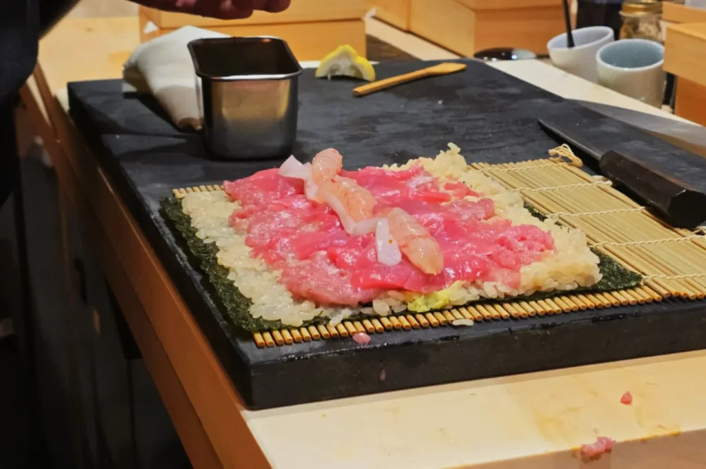 A sushi preparation scene showing a bamboo mat with nori, rice, and assorted raw fish toppings laid out before rolling.