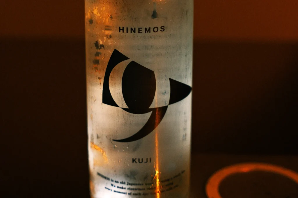 A close-up of a bottle of Hinemos Kuji sake with a minimalist black logo on a transparent label.