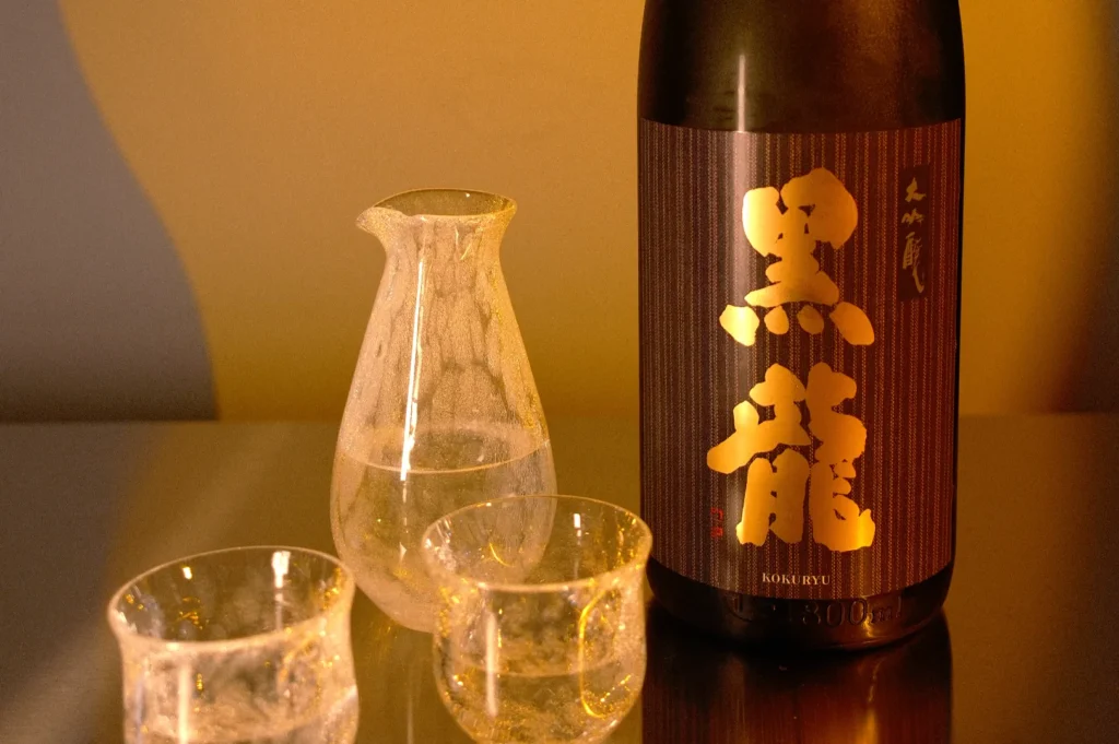 A bottle of sake with a decorative label, accompanied by a clear sake carafe and two small glasses, all set against a softly lit background.