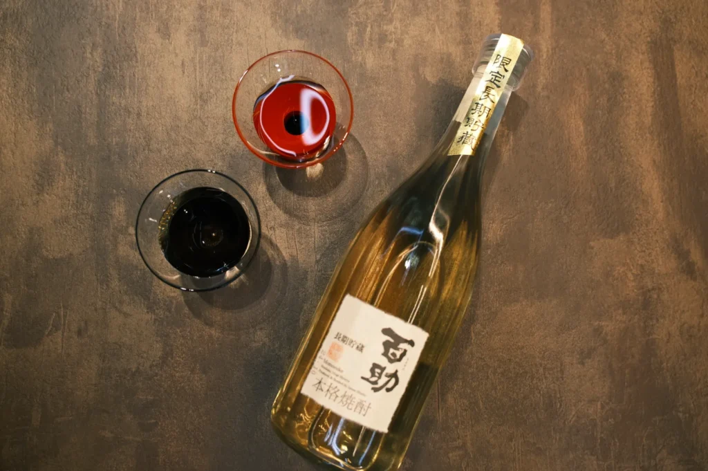 A bottle of sake with two glasses, one containing red liquid and the other containing dark liquid, on a textured surface.