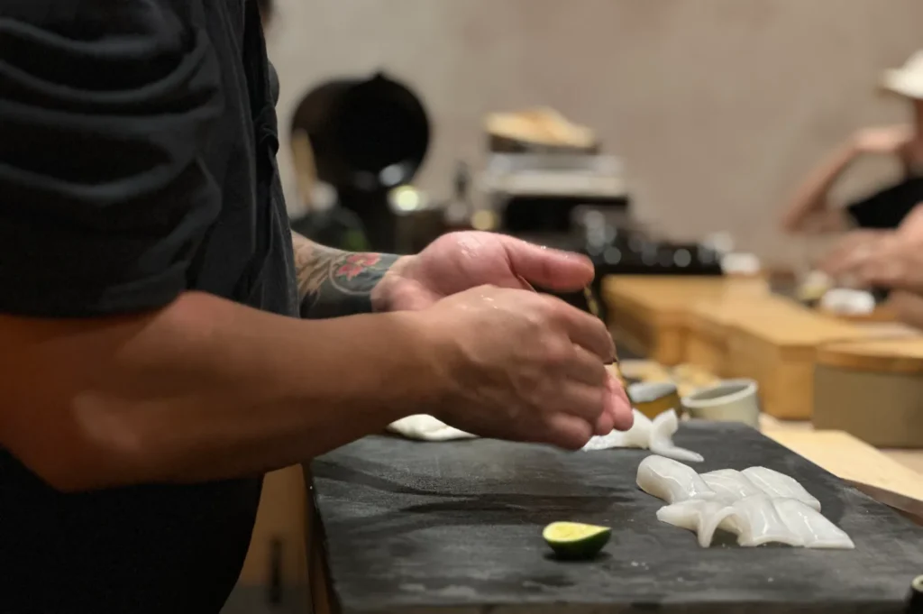 Chef's tattooed hands prepare sushi with precision on a dark cutting board. Fresh lime and sushi pieces are in focus, creating an artisanal vibe.