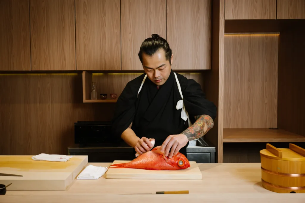 Chef Masa in a black kimono prepares a whole red fish on a wooden counter in a minimalist kitchen, conveying focus and tradition.