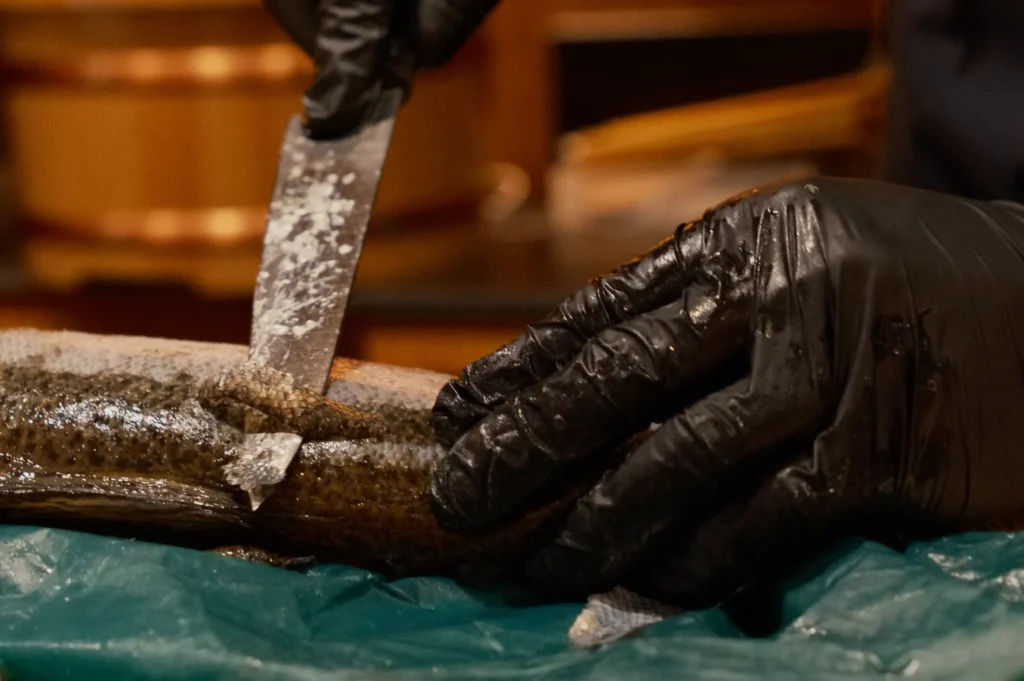 Chef Masa wearing black gloves is using a knife to fillet a fish on a blue cutting surface.