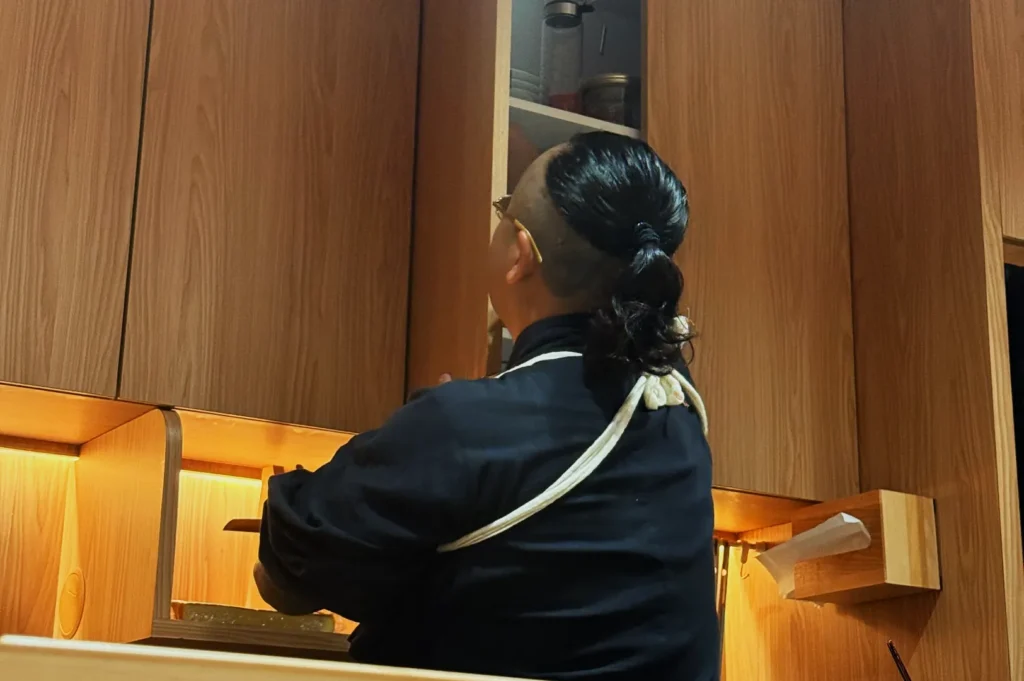 Chef Masa in a black uniform reaching up to a kitchen cabinet in a warm-toned wooden kitchen.