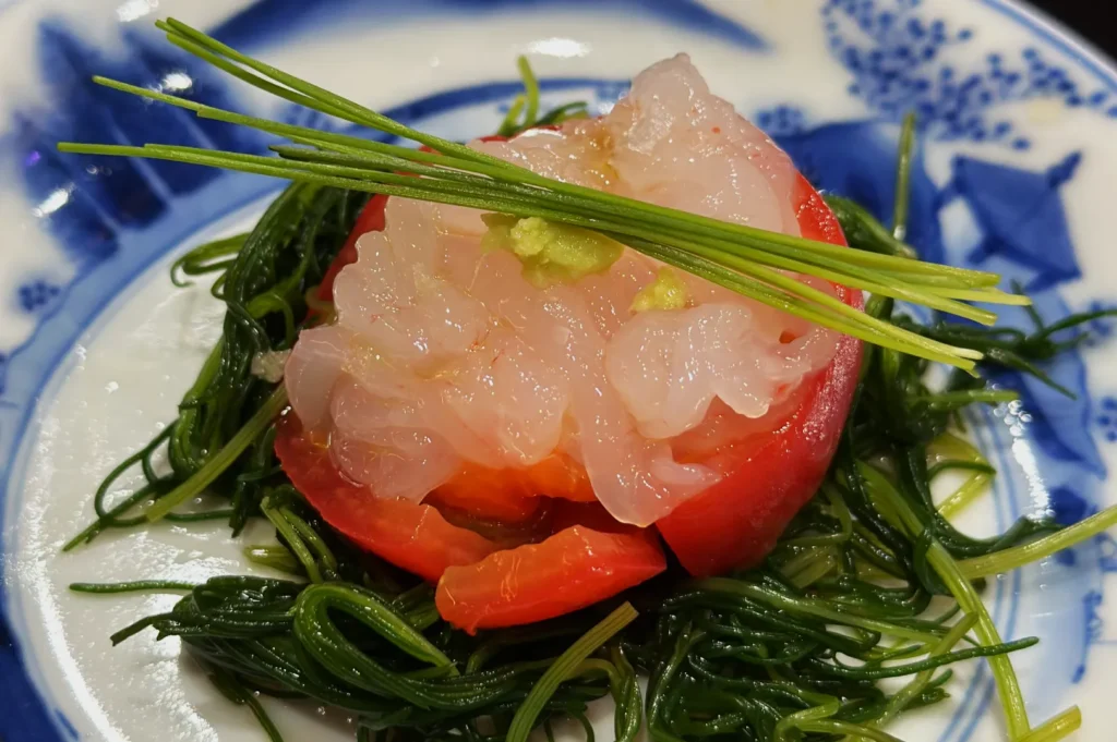 A dish featuring sashimi on tomato slices, garnished with green herbs, resting on a bed of seaweed. Served on a porcelain plate with a blue floral design.