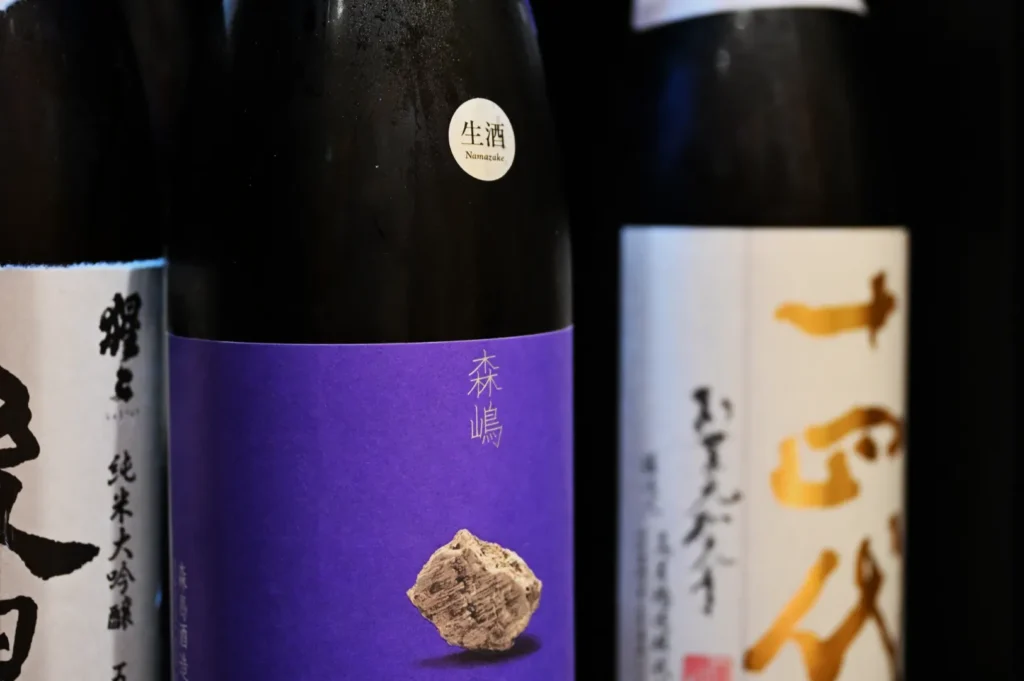 Three bottles of sake are displayed, featuring distinct labels. The bottle on the left has a white label with black text, the center bottle has a purple label with a rock-like shape in the foreground, and the right bottle has a white label with gold text.