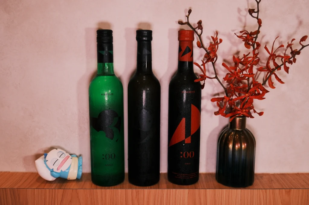 Three bottles of sake in different colors (green, black, and red) placed on a wooden shelf next to a decorative vase with red flowers and a small playful figurine.
