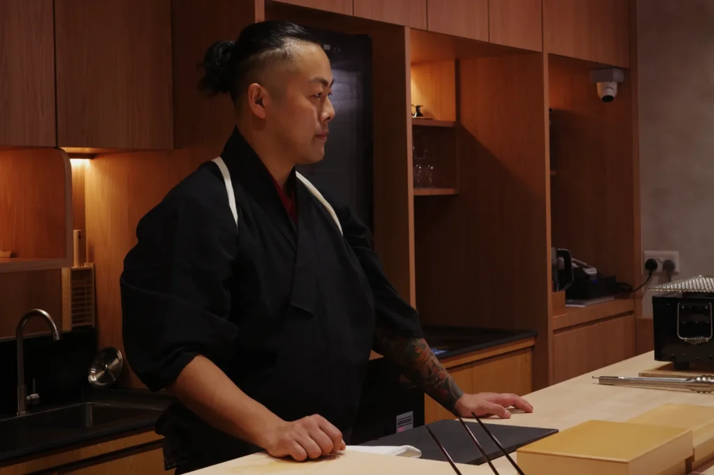 Chef Masa standing in a modern kitchen, wearing a traditional chef's jacket and looking thoughtfully at the countertop.