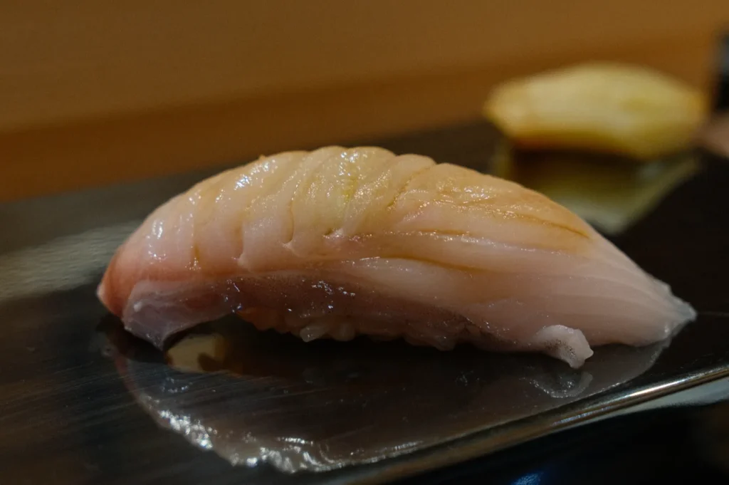 Close-up of a piece of sushi on a shiny metal plate, featuring a slice of raw fish with a glossy texture. A blurred piece of ginger is in the background.