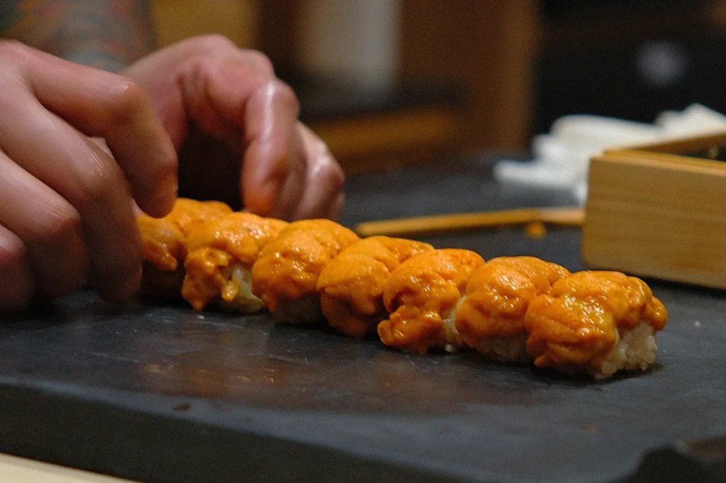 Chef Masa’s hands preparing sushi with orange seafood topping arranged on a slate surface.