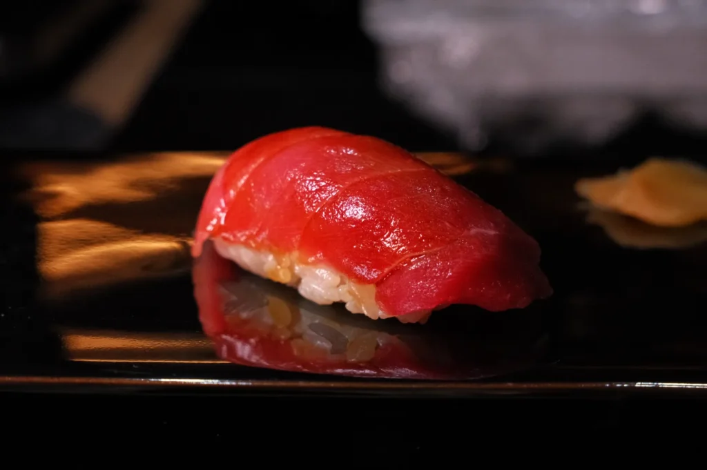A piece of sushi consisting of a slice of red fish (likely tuna) on top of a small mound of rice, served on a black plate.