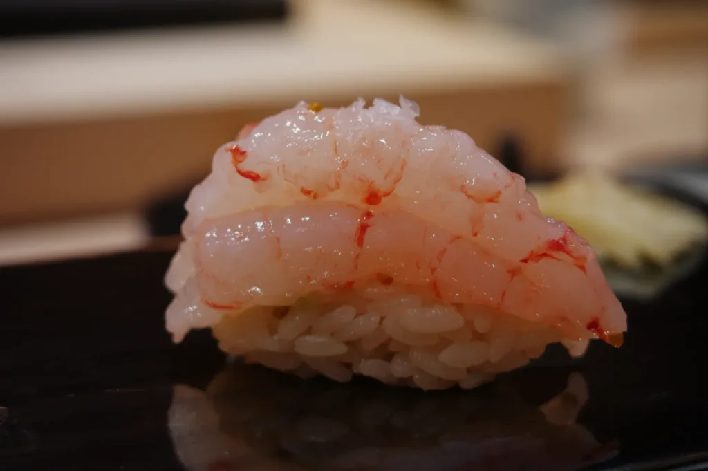 A piece of sushi with translucent pink shrimp on top of a mound of rice.