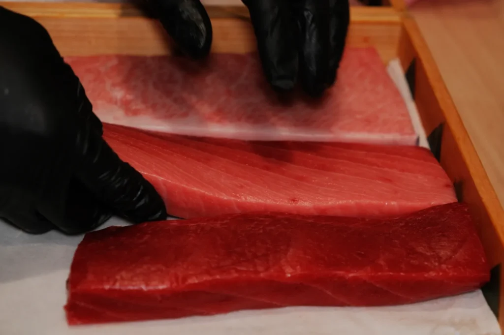 Hands in black gloves arrange three cuts of sushi-grade fish, varying in color from deep red to pale pink, on a wooden tray, suggesting freshness.
