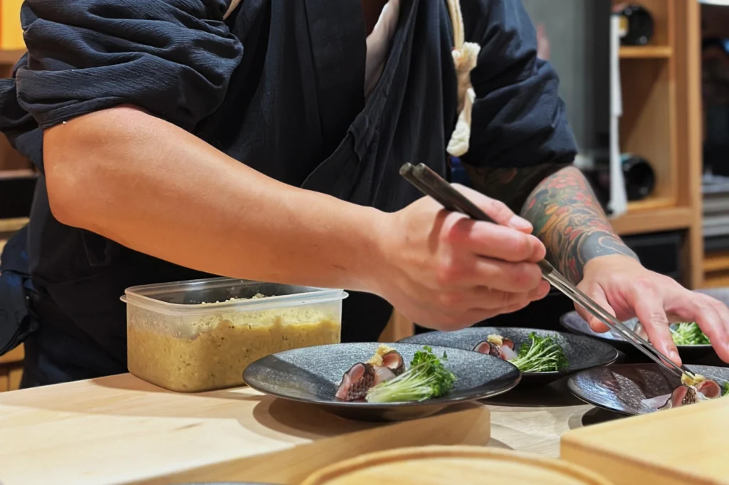 Chef Masa with tattooed arms skillfully plates sushi on black dishes, using chopsticks. Nearby, a container of food adds to the focused culinary scene.