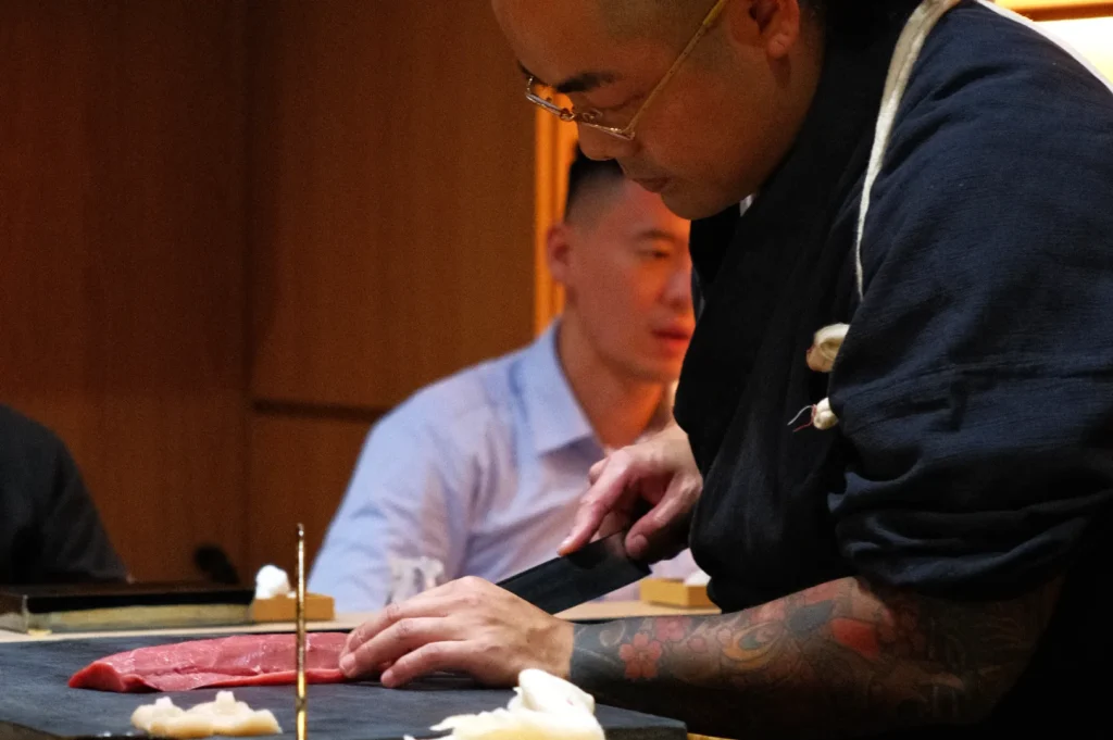 Chef Masa skillfully slicing fish in a restaurant setting, with a couple of patrons in the background.