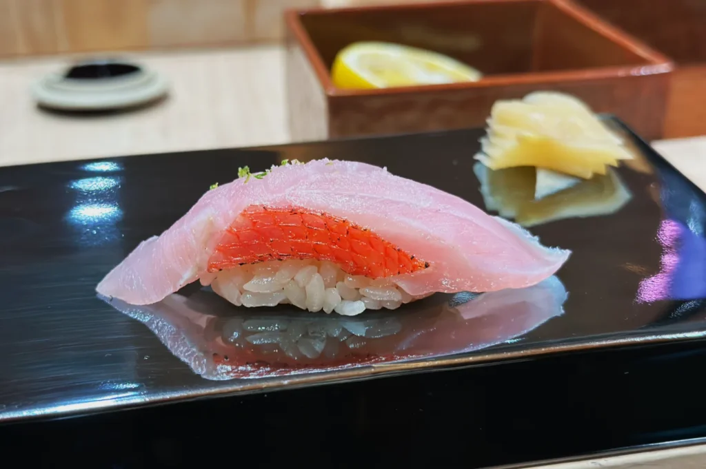 Close-up of nigiri sushi with a pink fish slice on rice, garnished with lime zest. Pickled ginger and lemon wedge are blurred in the background.