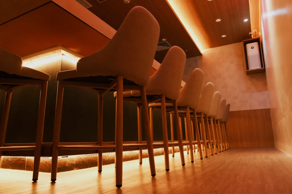 A low-angle view of a row of modern wooden chairs with soft seats, positioned in a dimly lit interior space featuring warm lighting.