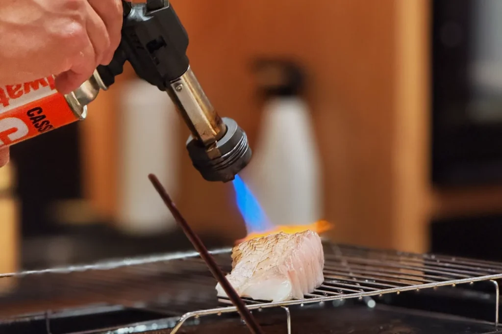 A hand using a kitchen torch to sear a piece of fish on a grill rack.