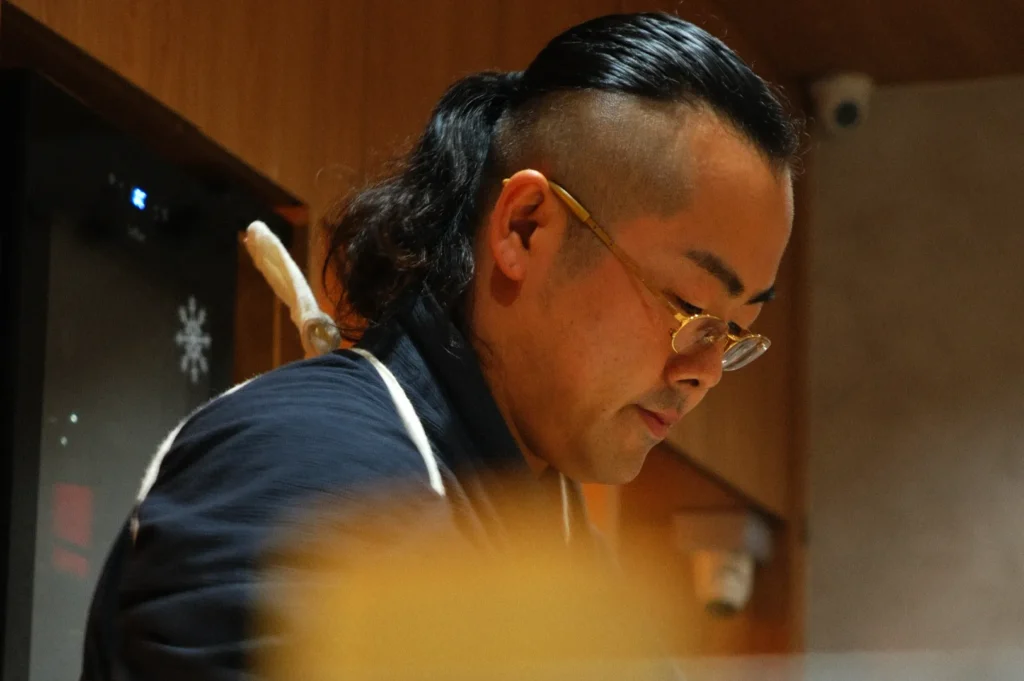 Chef Masa with glasses and a ponytail is focused on preparing food in a restaurant kitchen.