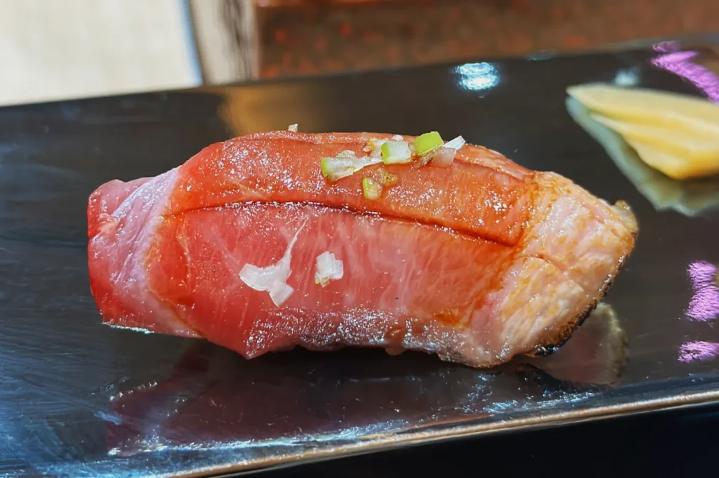 Close-up of a piece of sushi on a black plate. The sushi features thick slices of marbled pink fish topped with diced green onions, exuding a fresh, appetizing look.