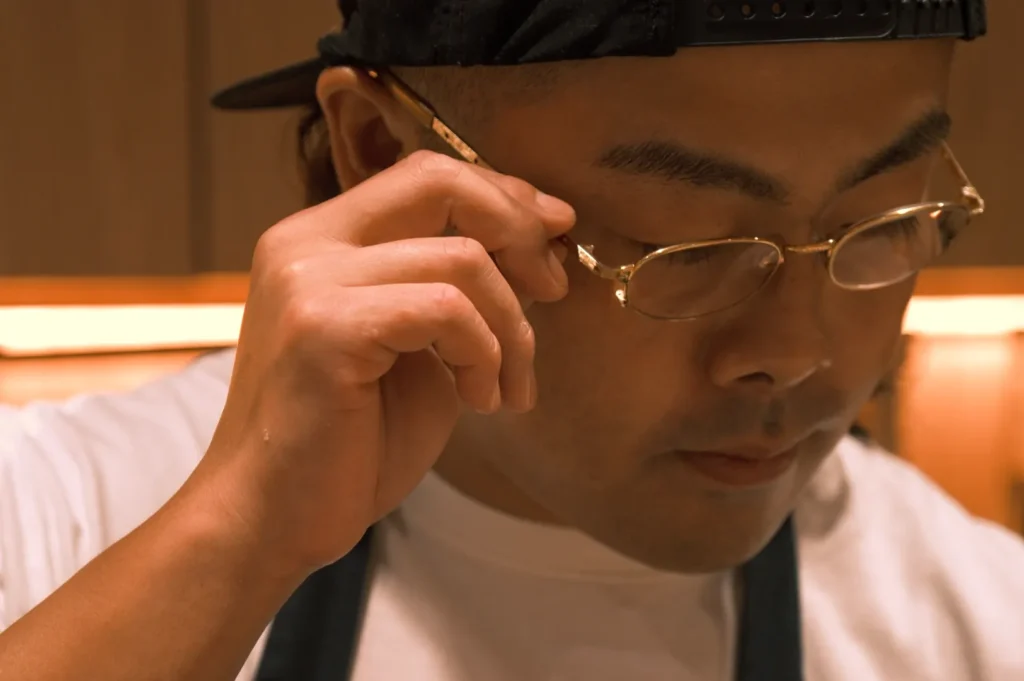 A close-up of Chef Masa adjusting his glasses, wearing a cap and a shirt, with a focused expression.
