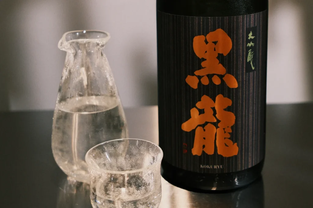 A bottle of sake with a distinctive black and orange label next to a glass and a carafe filled with clear liquid.
