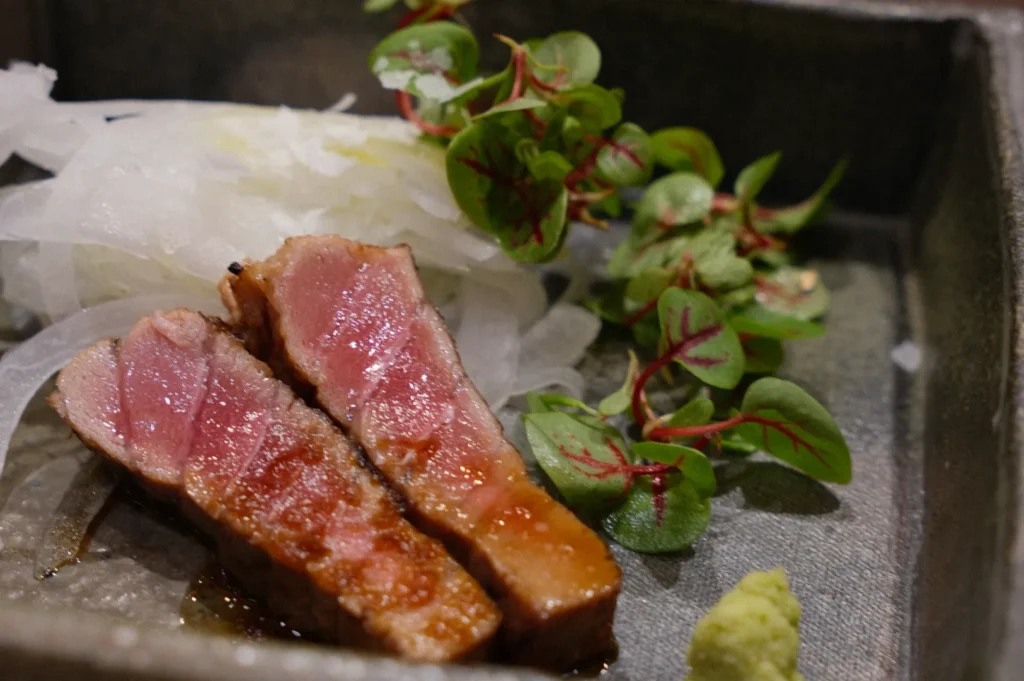 Sliced seared beef with a pink center, garnished with fresh greens and shredded white radish, resting on a square, dark ceramic plate. A dollop of wasabi adds color.