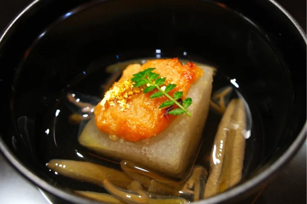 A dish featuring a piece of fish or seafood on a square block of tofu or a root vegetable, garnished with a small green leaf and gold leaf, served in a clear broth with slivers of something resembling mushrooms or root vegetables.