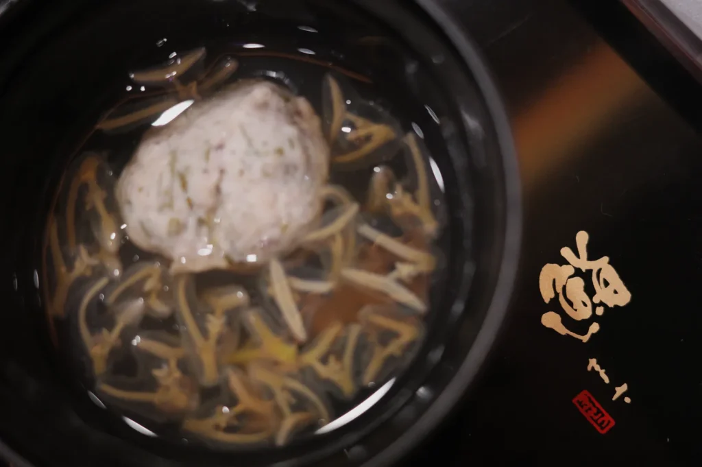A bowl of soup containing a large meatball and strips of vegetables, with a decorative symbol on the side.