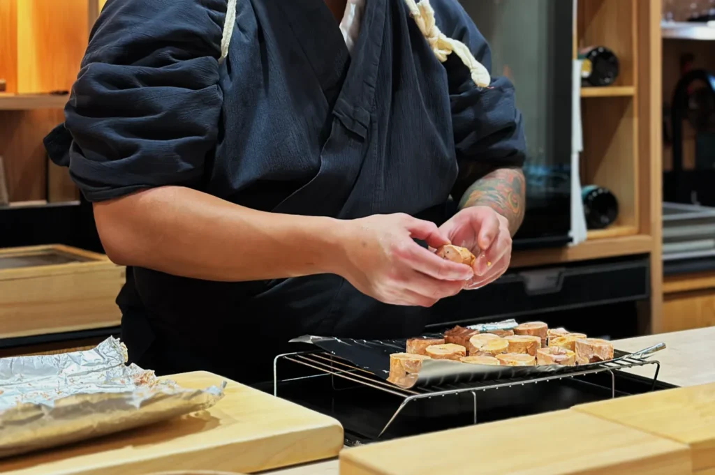 Chef in a dark robe prepares food in a modern kitchen, arranging sliced ingredients on a tray. The scene conveys focus and culinary expertise.