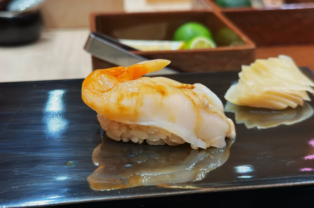 A piece of sushi topped with a glossy, orange-tinted shrimp on a black plate. Beside it, a small serving of thinly sliced ginger. Background shows blurred lime.