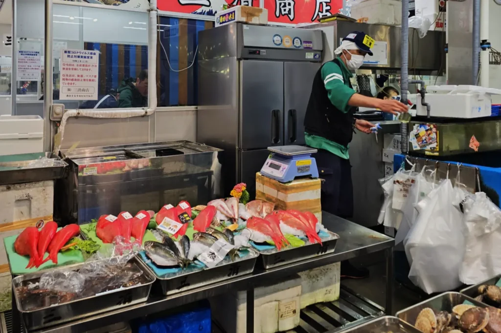 Market scene with a fish vendor preparing seafood. Fresh fish varieties displayed on trays in the foreground. Vendor in uniform and mask stands nearby.