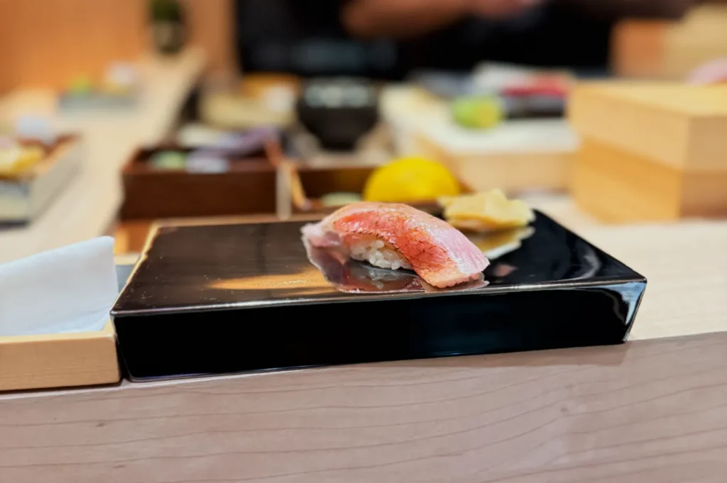 A piece of sushi on a black plate, with other sushi and garnishes in the background.