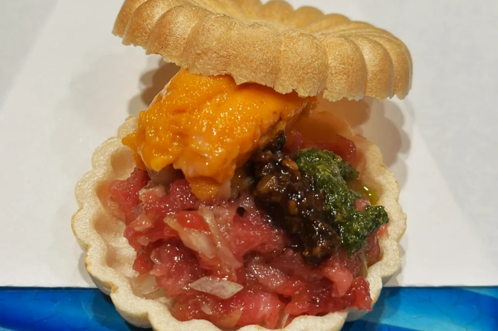 Open-faced tart with a flaky shell, filled with vibrant orange sea urchin, marinated raw fish, and green garnish, creating a fresh, gourmet presentation.