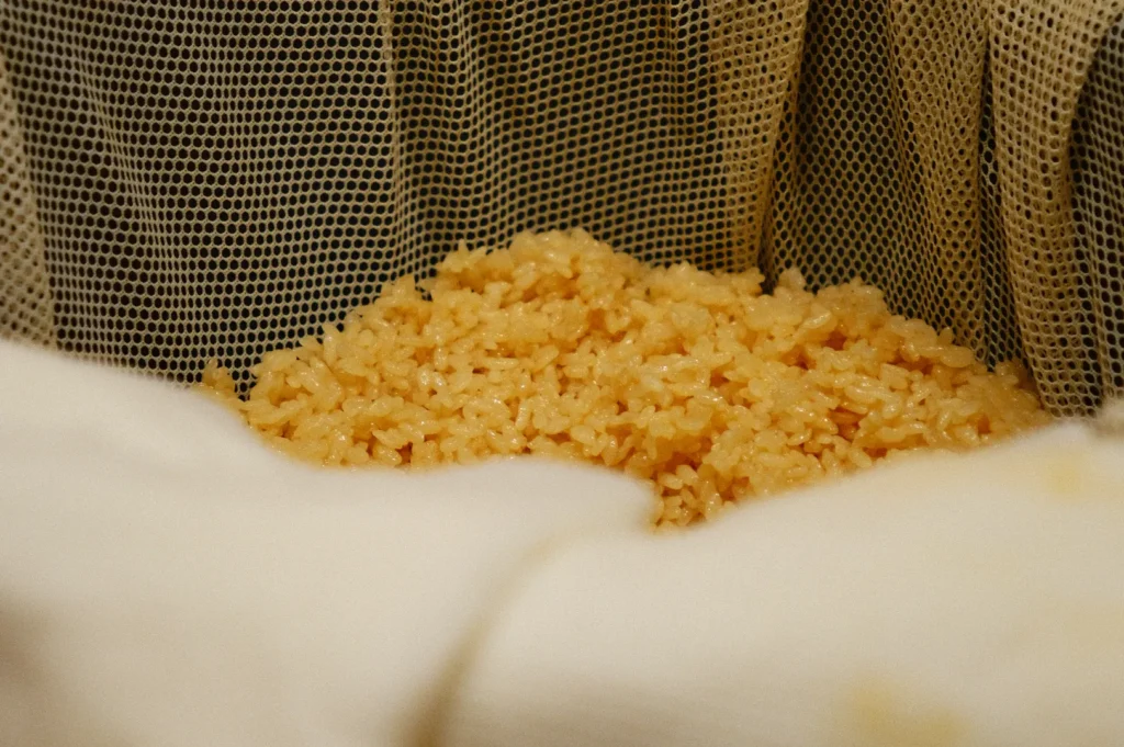 Close-up of a mound of golden rice on a netted cloth, with soft lighting creating a warm, cozy atmosphere. Two white-gloved hands gently hold the cloth edges.