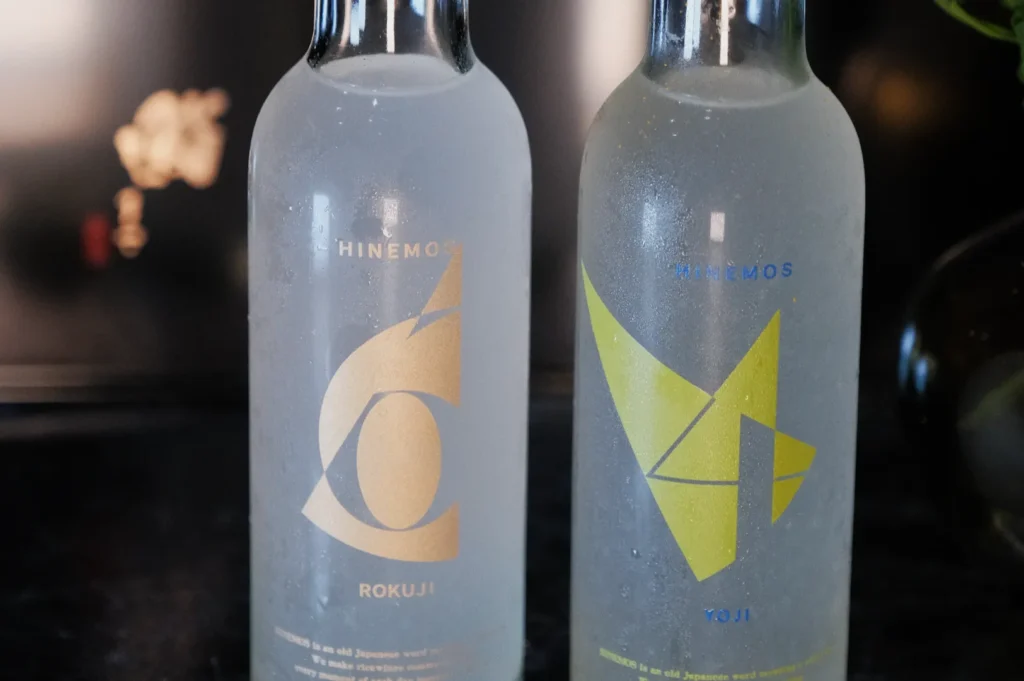 Two glass bottles of sake with distinctive labels; one features a golden "6" and the word "Rokku," while the other has a yellow "4" and the word "Toni." The bottles appear chilled with condensation on the surface.