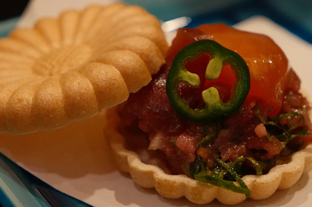 A delicate open tart filled with rich tuna tartare, garnished with a vibrant jalapeño slice and a glossy sauce, resting on a floral-patterned pastry.