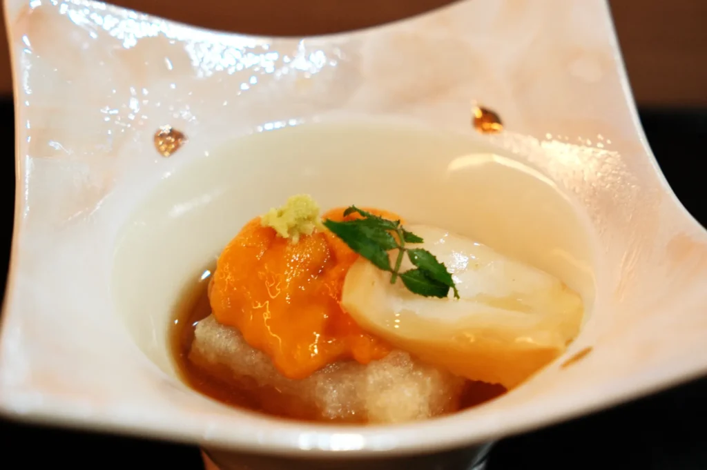 A small dish containing a creamy substance topped with orange sea urchin, a slice of white seafood, garnished with a sprig of green herb and a small amount of grated wasabi.
