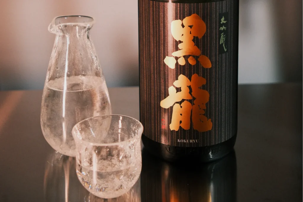 A bottle of sake with kanji characters, accompanied by a small cup and a glass pitcher filled with clear liquid.