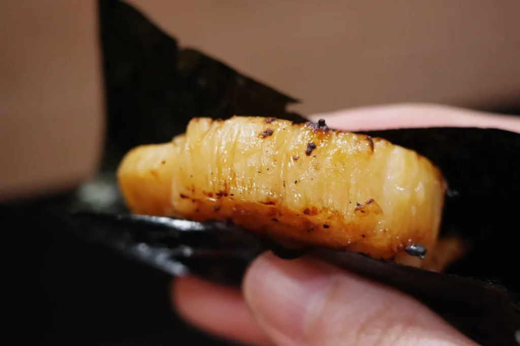 A piece of grilled fish wrapped in a seaweed leaf being held by a hand.