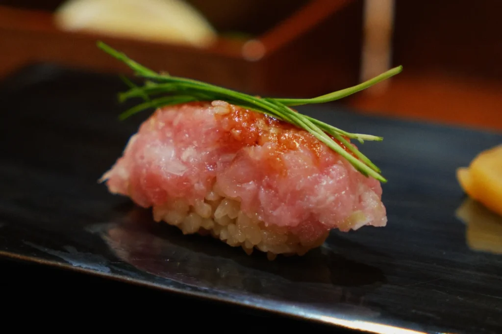 Close-up of a piece of sushi with pink minced fish on rice, garnished with green chives, on a dark plate. The scene suggests fine dining.