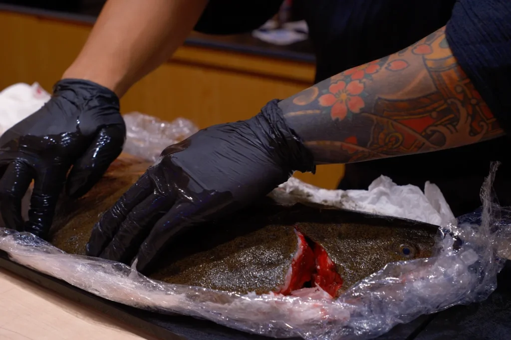 Tattooed arm and gloved hands preparing a fish for cooking. The fish is partially wrapped in clear plastic, revealing its textured skin and deep cut.