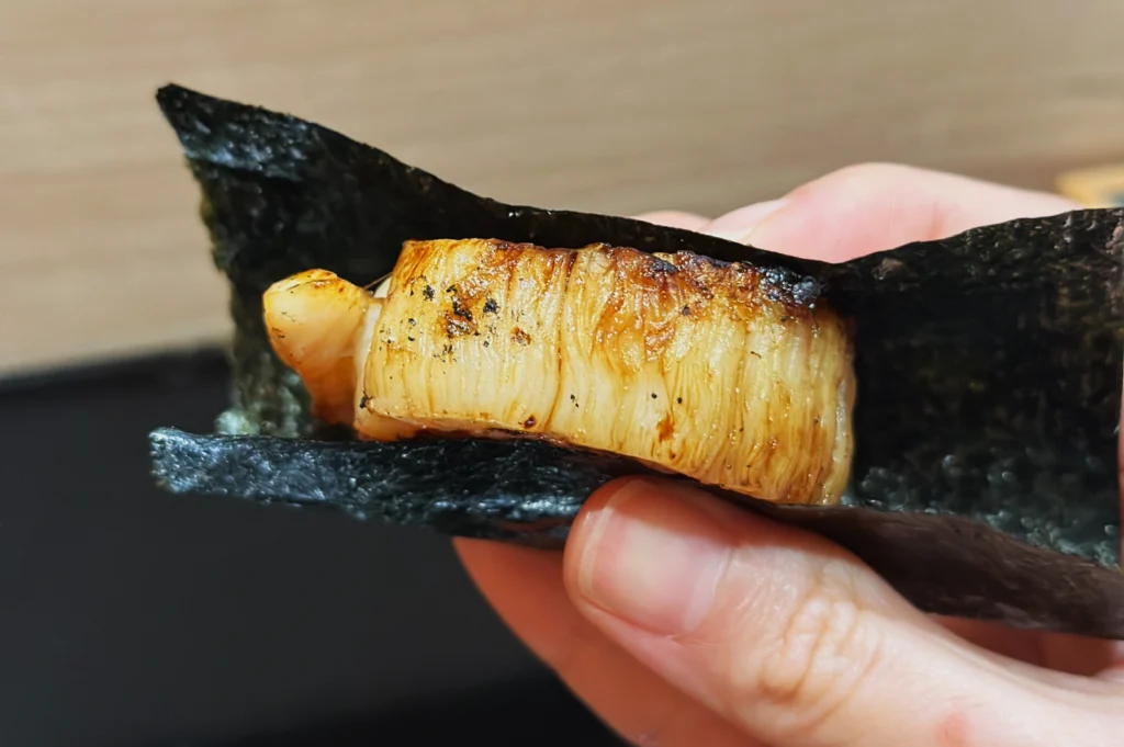 Close-up of a hand holding a grilled scallop encased in a folded piece of dark green seaweed. The scallop is golden-brown, enhancing its savory appeal.