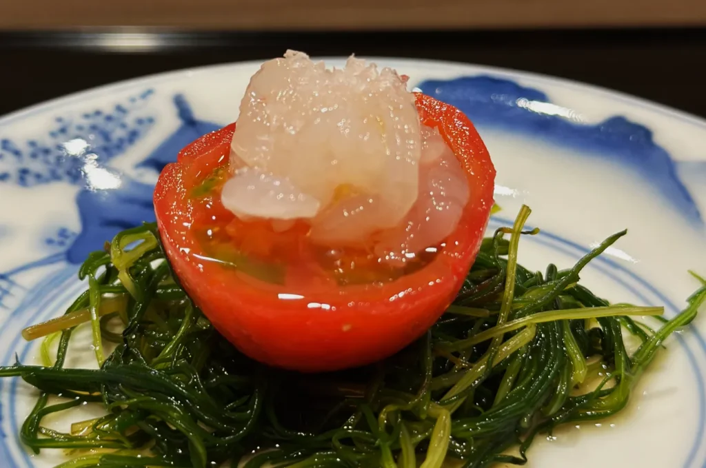 A halved tomato filled with a translucent topping sits on a bed of fresh green seaweed. The dish is on a white and blue plate, creating a refreshing, elegant presentation.