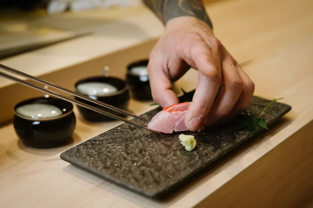Chef Masa’s hand with a tattoo prepares sushi with chopsticks on a dark rectangular plate, garnished with wasabi. Three soy sauce bowls are blurred in the background.