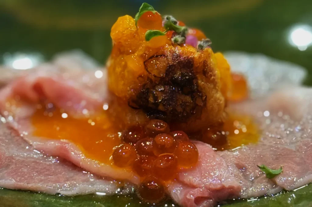 A close-up image of a gourmet dish featuring thin slices of meat topped with vibrant orange and yellow elements, including possibly fish roe and a golden crispy item, all arranged artistically on a plate.