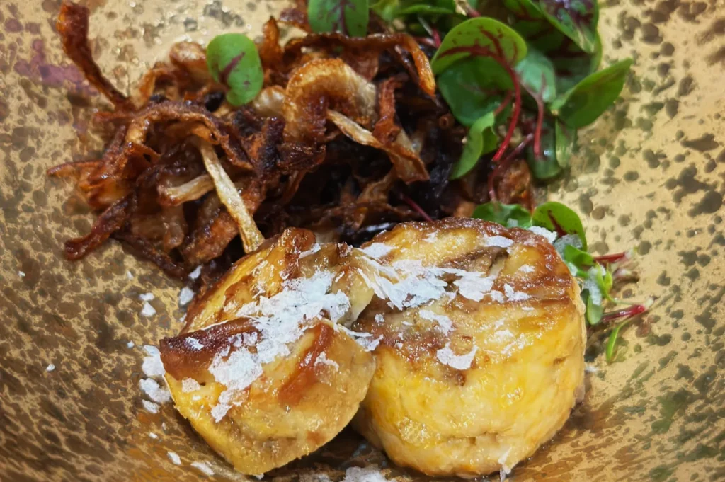 Two golden-brown, flakey pastries topped with sea salt, served on a speckled plate with crispy fried onions and fresh green herbs, creating a savory and appetizing look.