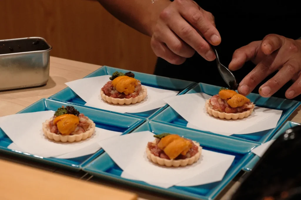 Hands carefully spoon sea urchin onto tartlets with tuna tartare on blue plates, creating an elegant and gourmet presentation.